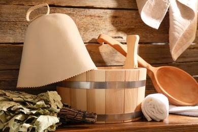 Photo of Sauna equipment. Bucket, ladle, felt wool hat, oak whisk and towels on wooden table indoors