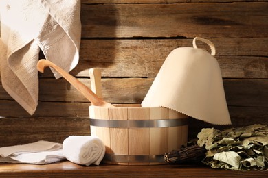 Photo of Sauna equipment. Bucket with ladle, felt wool hat, oak whisk and towels on wooden table indoors