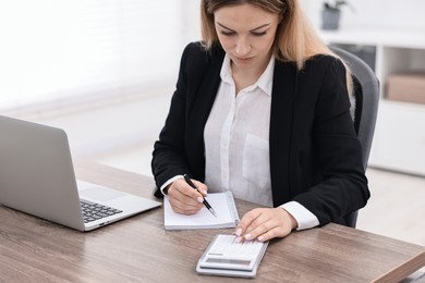 Photo of Banker with notebook using calculator at wooden table in office