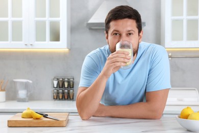 Handsome man drinking water with lemon at white marble table in kitchen