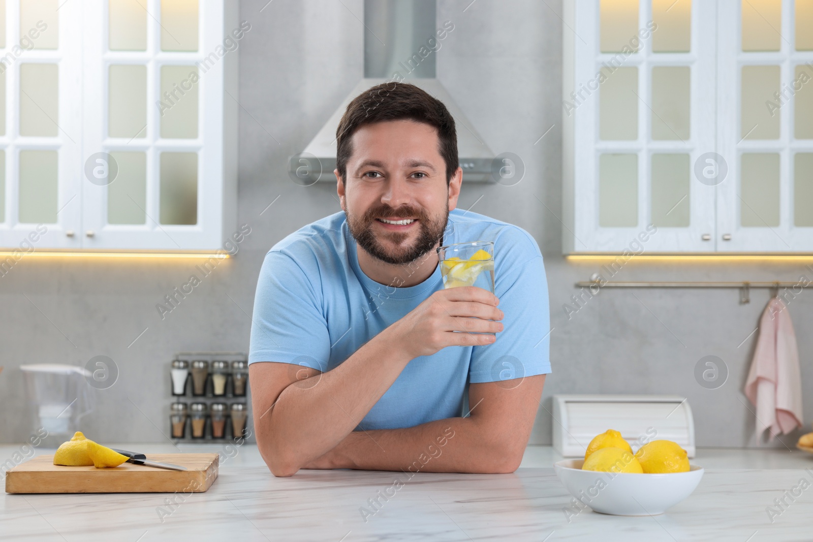 Photo of Happy man holding glass of water with lemon at white marble table in kitchen