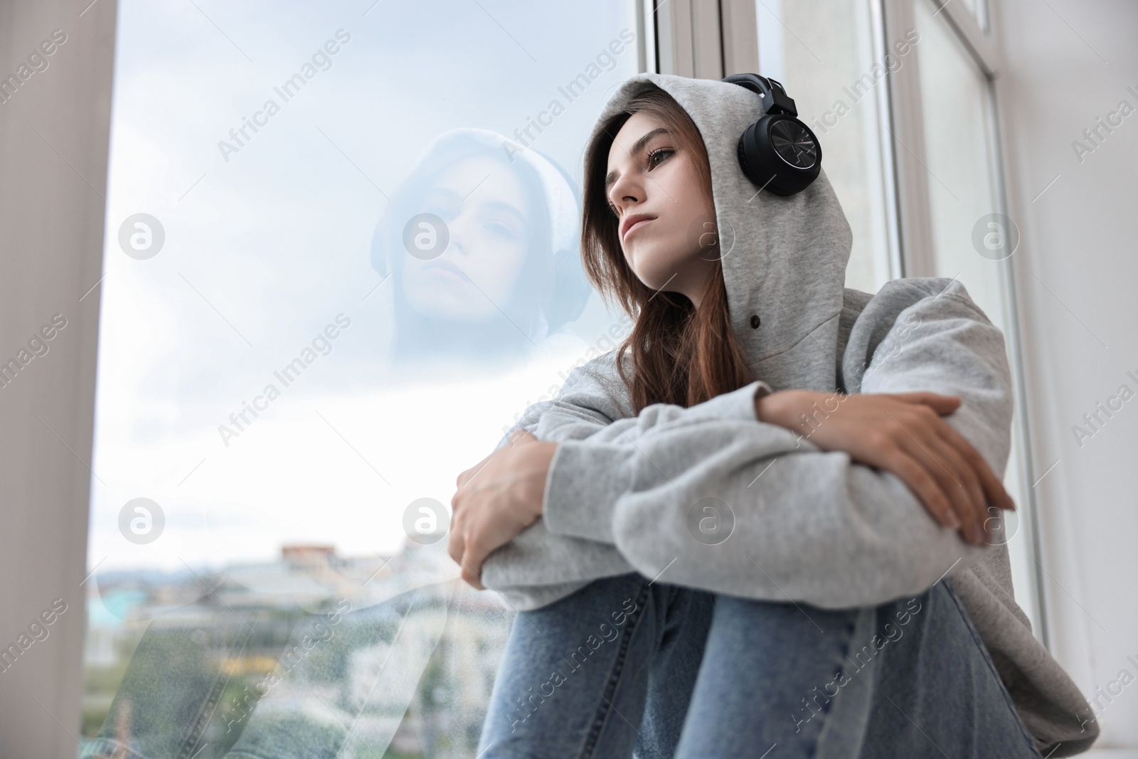 Photo of Loneliness concept. Sad teenage girl in headphones listening to music near window at home, low angle view