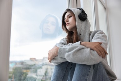 Loneliness concept. Sad teenage girl in headphones listening to music near window at home, low angle view