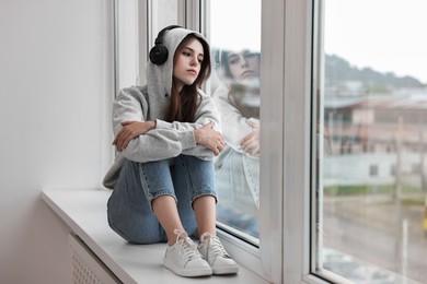 Loneliness concept. Sad teenage girl in headphones listening to music on windowsill at home