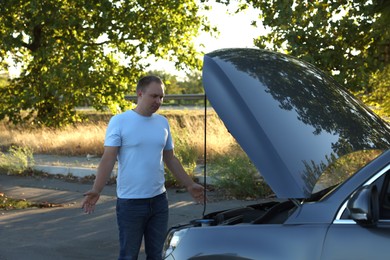 Stressed man looking under hood of broken car outdoors