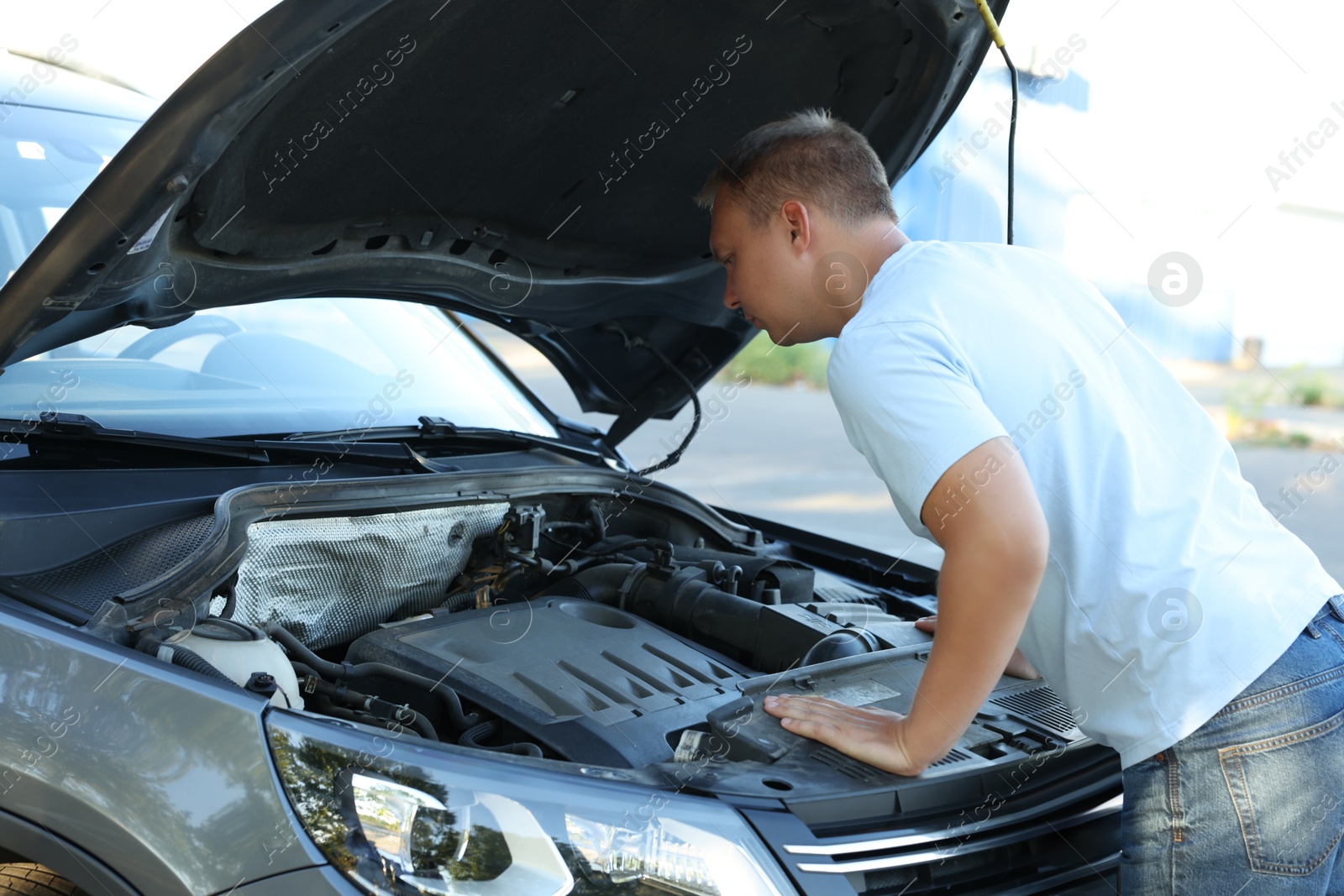 Photo of Stressed man looking under hood of broken car outdoors