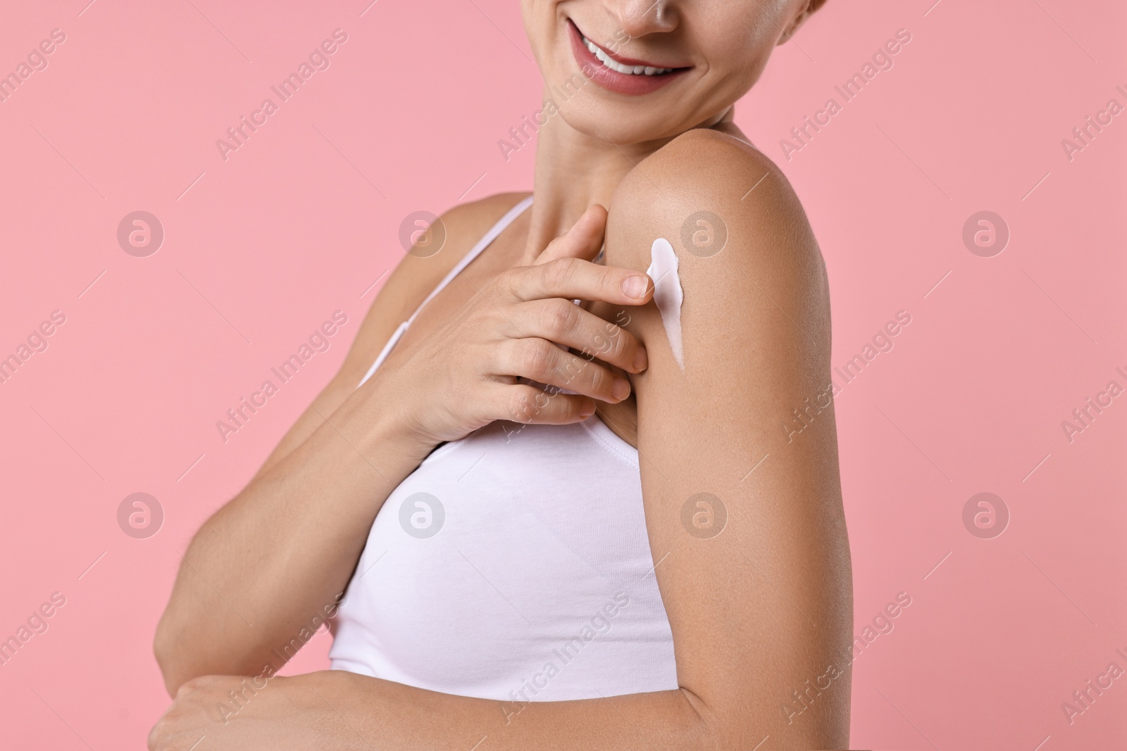 Photo of Smiling woman applying cream onto shoulder against pink background, closeup. Body care