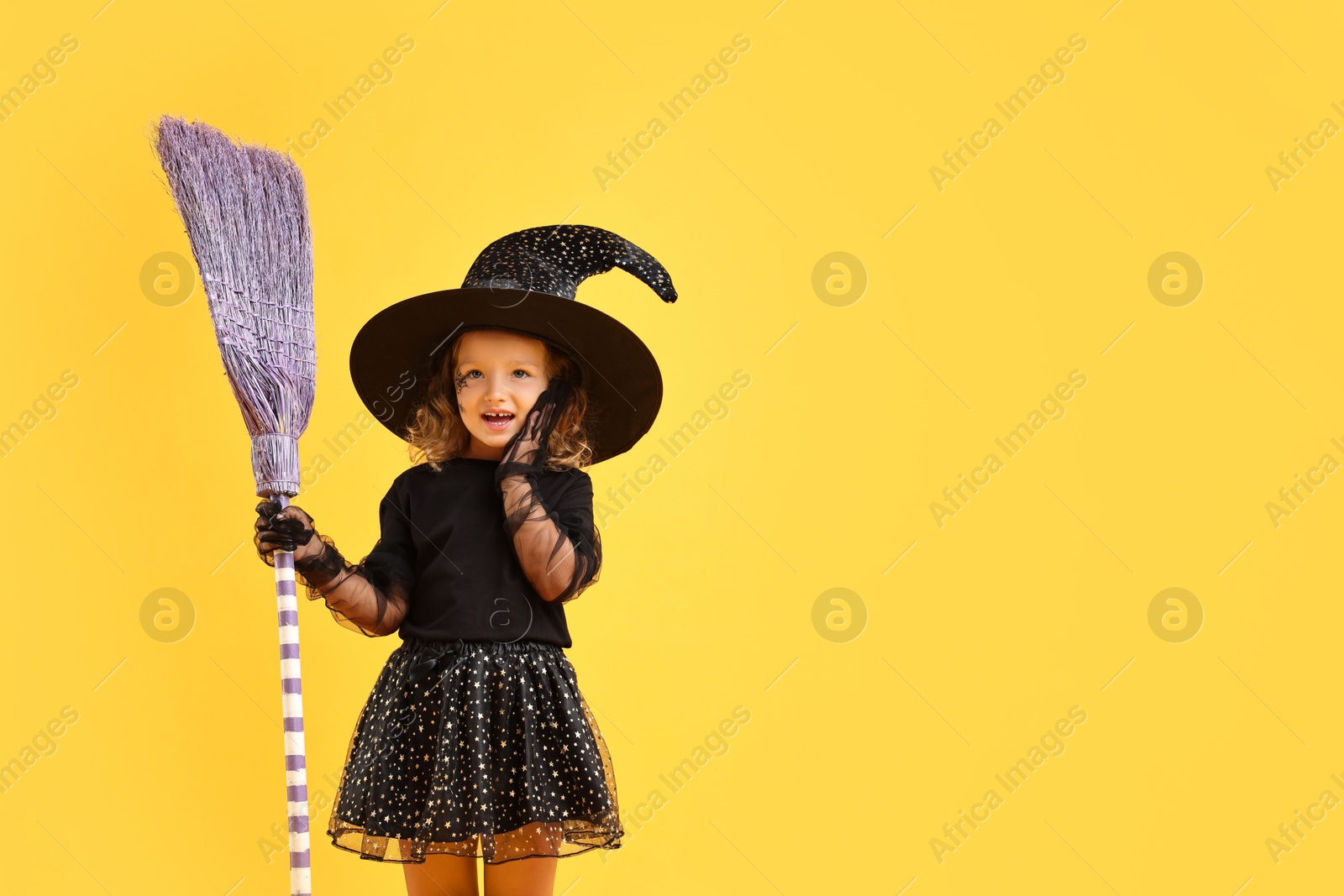 Photo of Cute girl with broom dressed like witch on yellow background, space for text. Halloween celebration