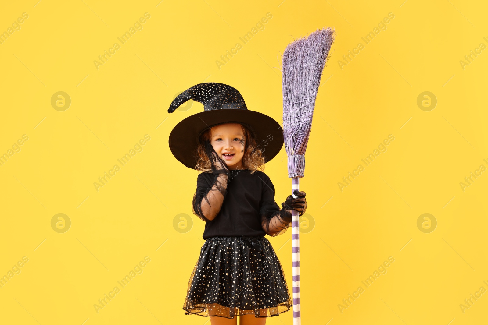 Photo of Cute girl with broom dressed like witch on yellow background. Halloween celebration