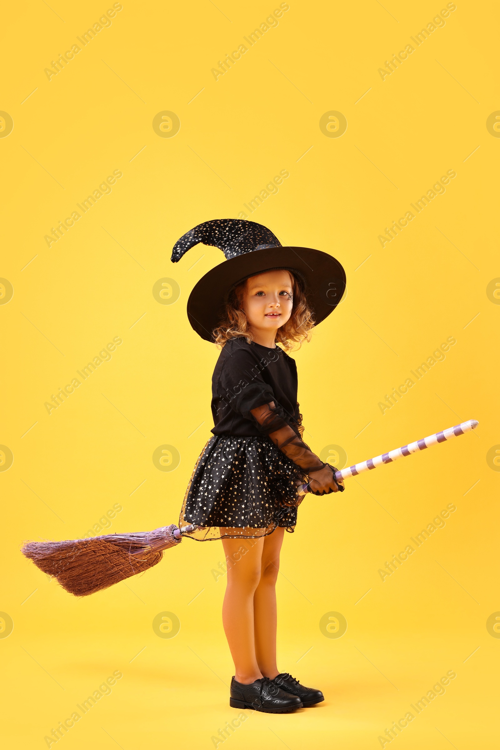 Photo of Cute girl with broom dressed like witch on yellow background. Halloween celebration