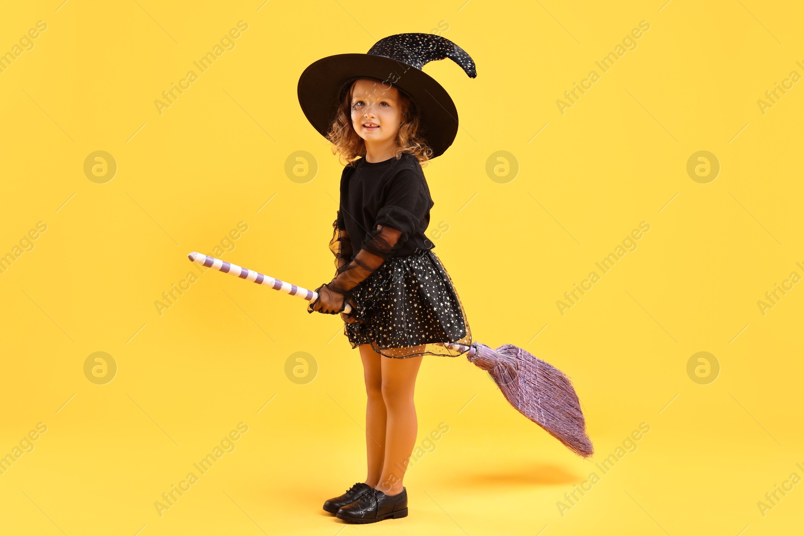 Photo of Cute girl with broom dressed like witch on yellow background. Halloween celebration