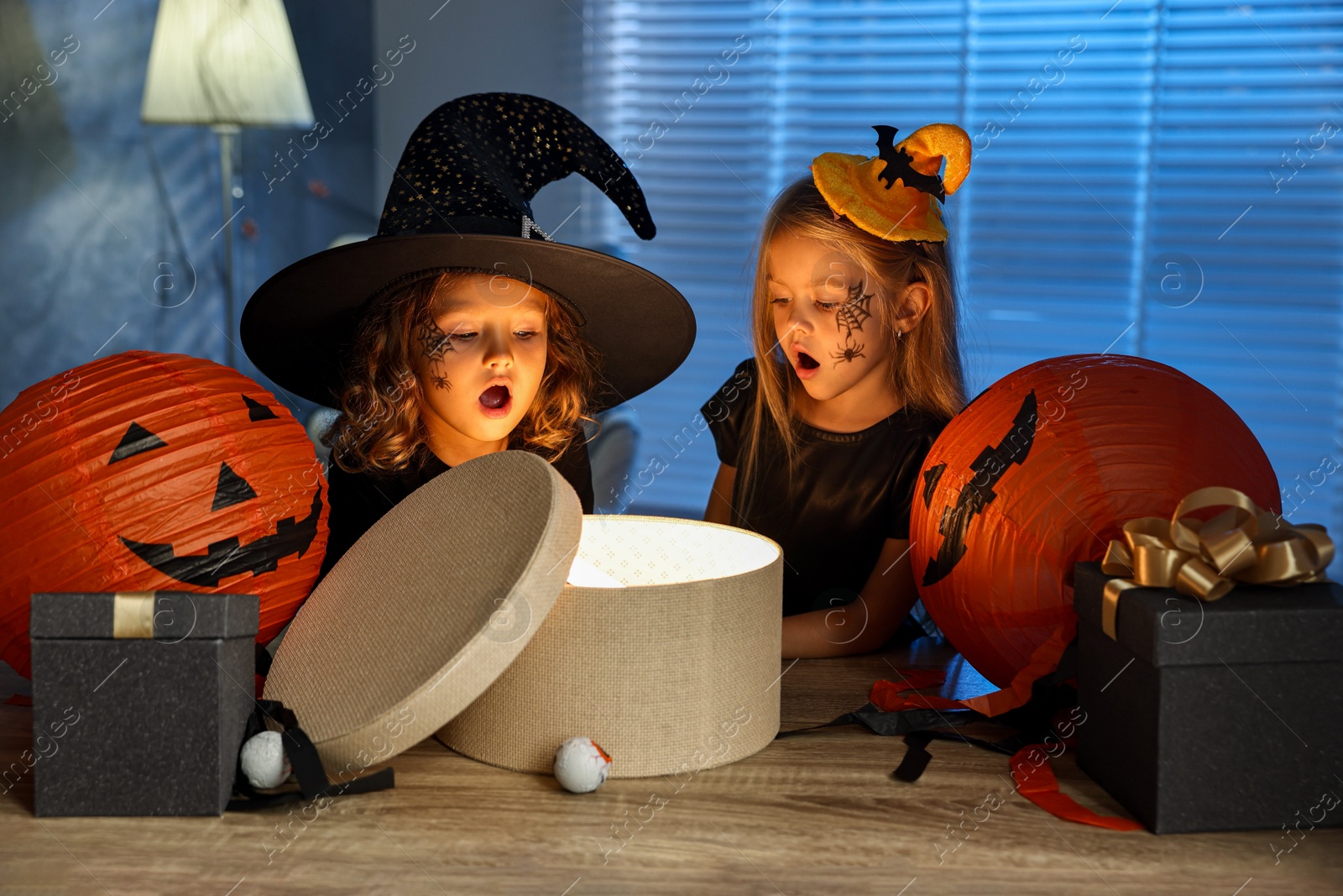 Photo of Surprised children in costumes with festive decor and gift boxes indoors at night. Halloween celebration