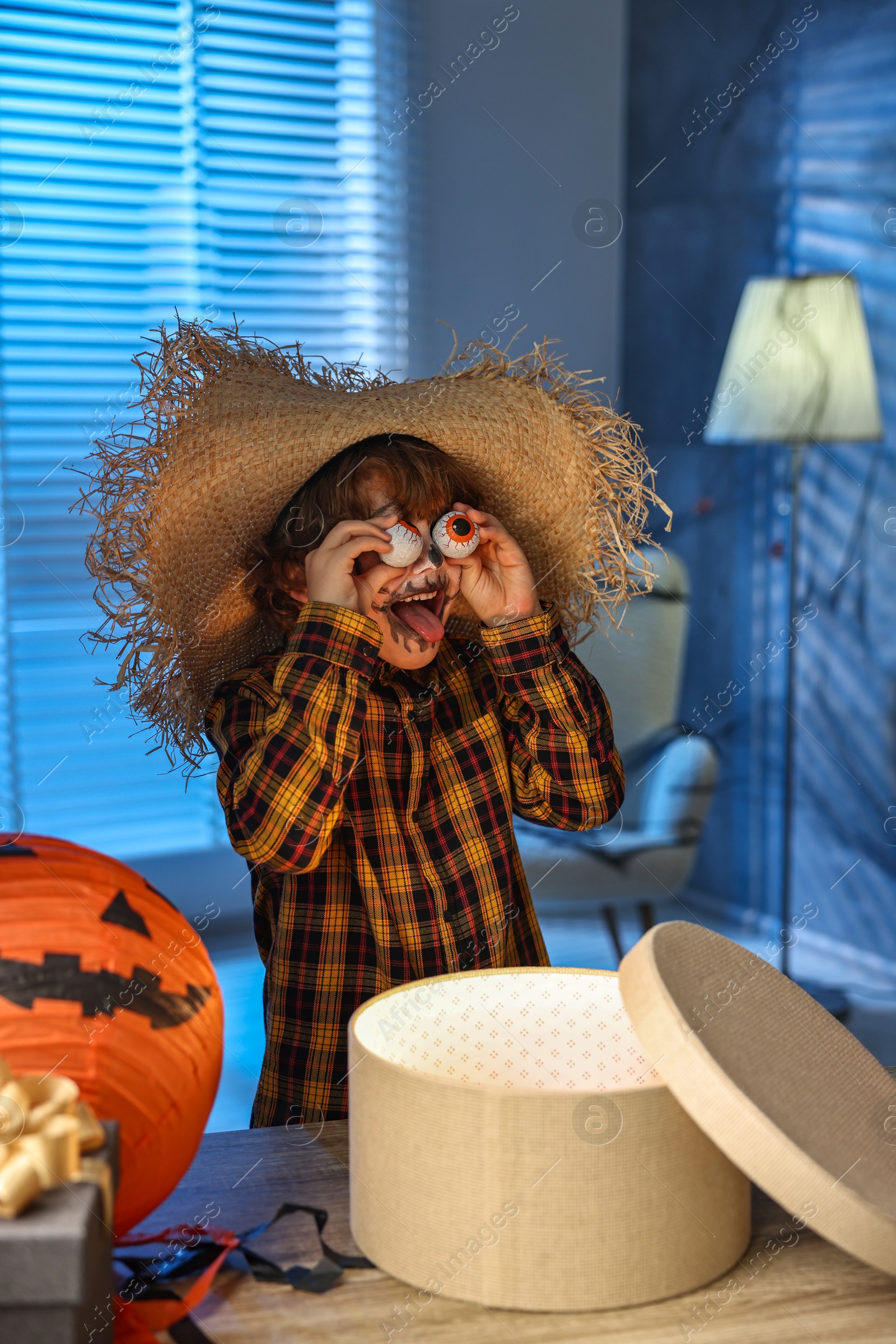 Photo of Cute boy dressed like scarecrow with decorative eyeballs, festive decor and gift boxes indoors at night. Halloween celebration