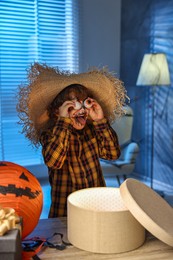 Photo of Cute boy dressed like scarecrow with decorative eyeballs, festive decor and gift boxes indoors at night. Halloween celebration