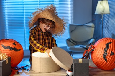 Cute boy dressed like scarecrow with festive decor and gift boxes indoors at night. Halloween celebration