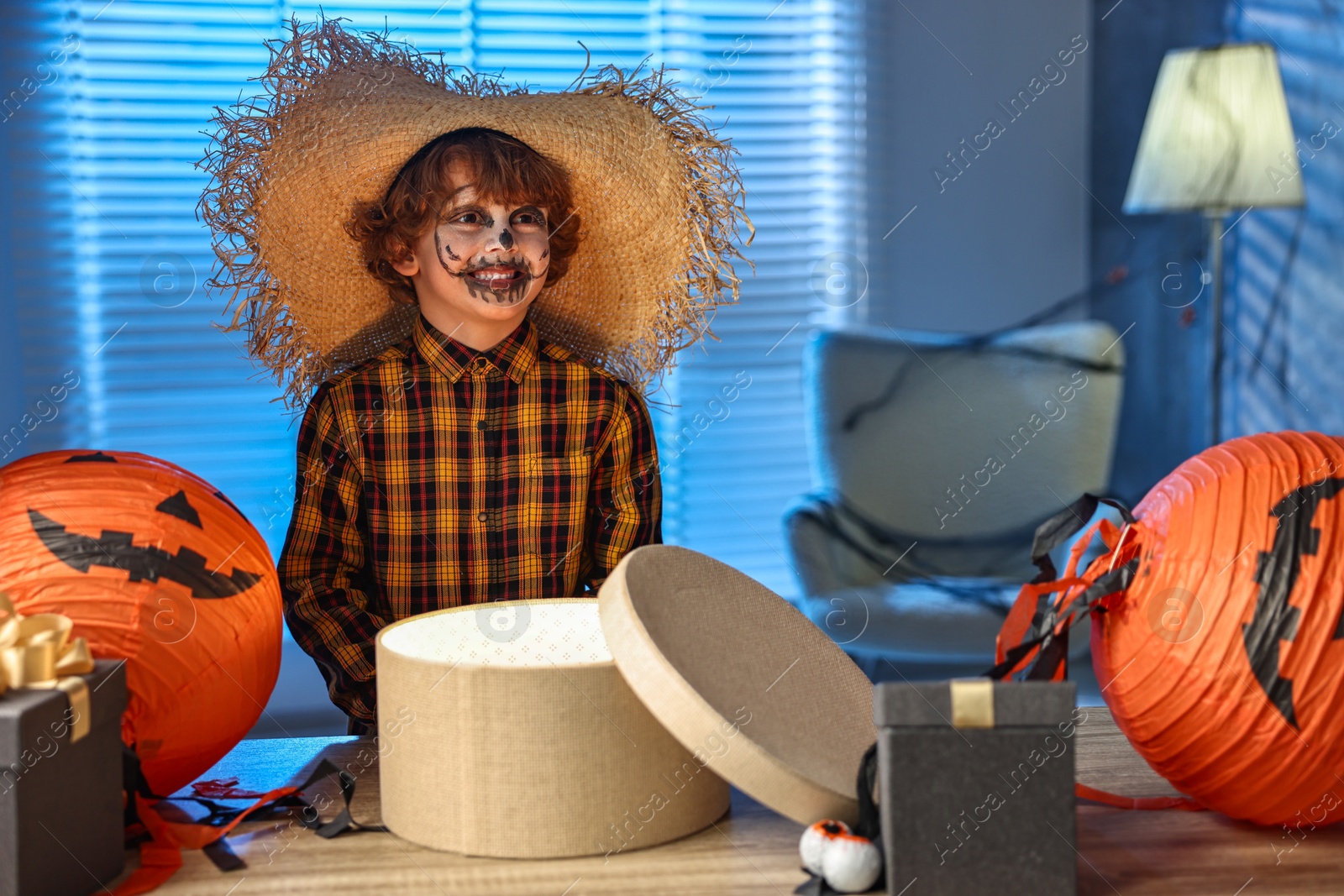 Photo of Cute boy dressed like scarecrow with festive decor and gift boxes indoors at night. Halloween celebration