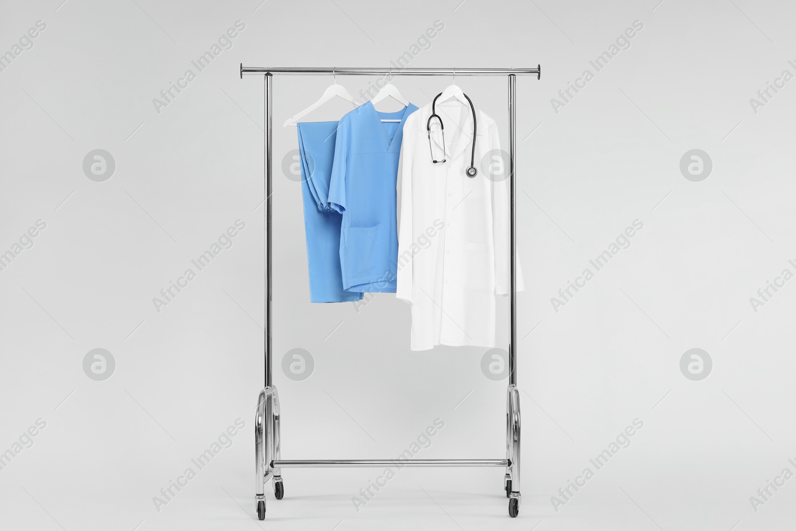 Photo of Different medical workers' uniforms and stethoscope on clothing rack against white background