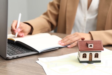 Photo of House hunting. Woman with laptop, papers, notebook and house figure at wooden table, closeup