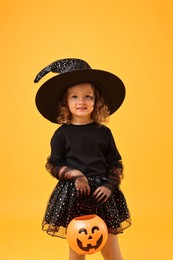 Photo of Cute girl with pumpkin bucket dressed like witch on yellow background. Halloween celebration