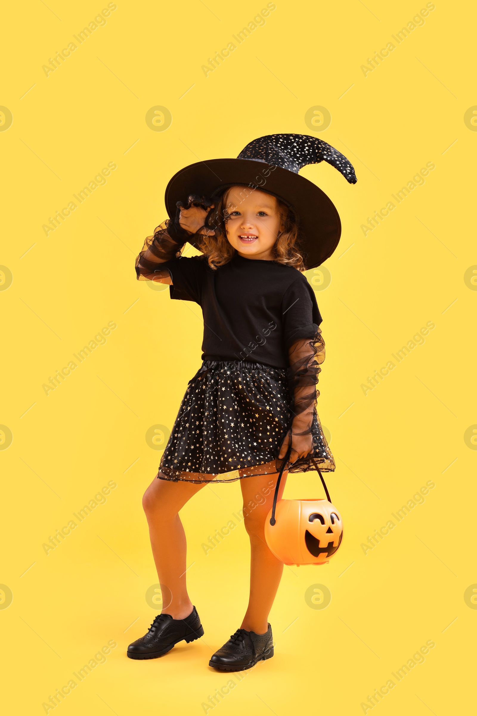 Photo of Cute girl with pumpkin bucket dressed like witch on yellow background. Halloween celebration