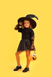 Photo of Cute girl with pumpkin bucket dressed like witch on yellow background. Halloween celebration