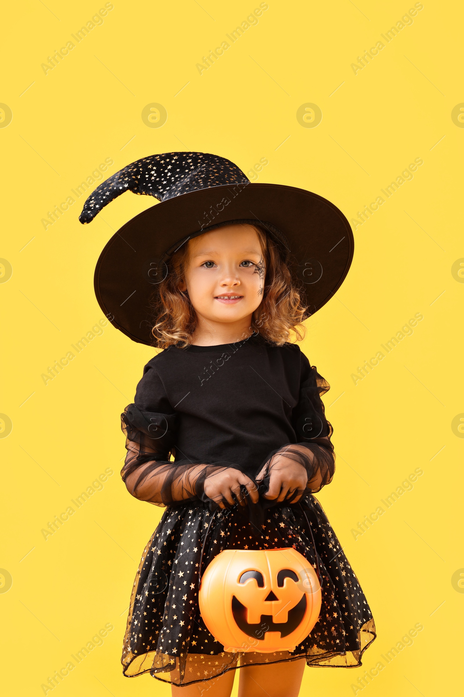 Photo of Cute girl with pumpkin bucket dressed like witch on yellow background. Halloween celebration