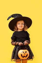 Cute girl with pumpkin bucket dressed like witch on yellow background. Halloween celebration