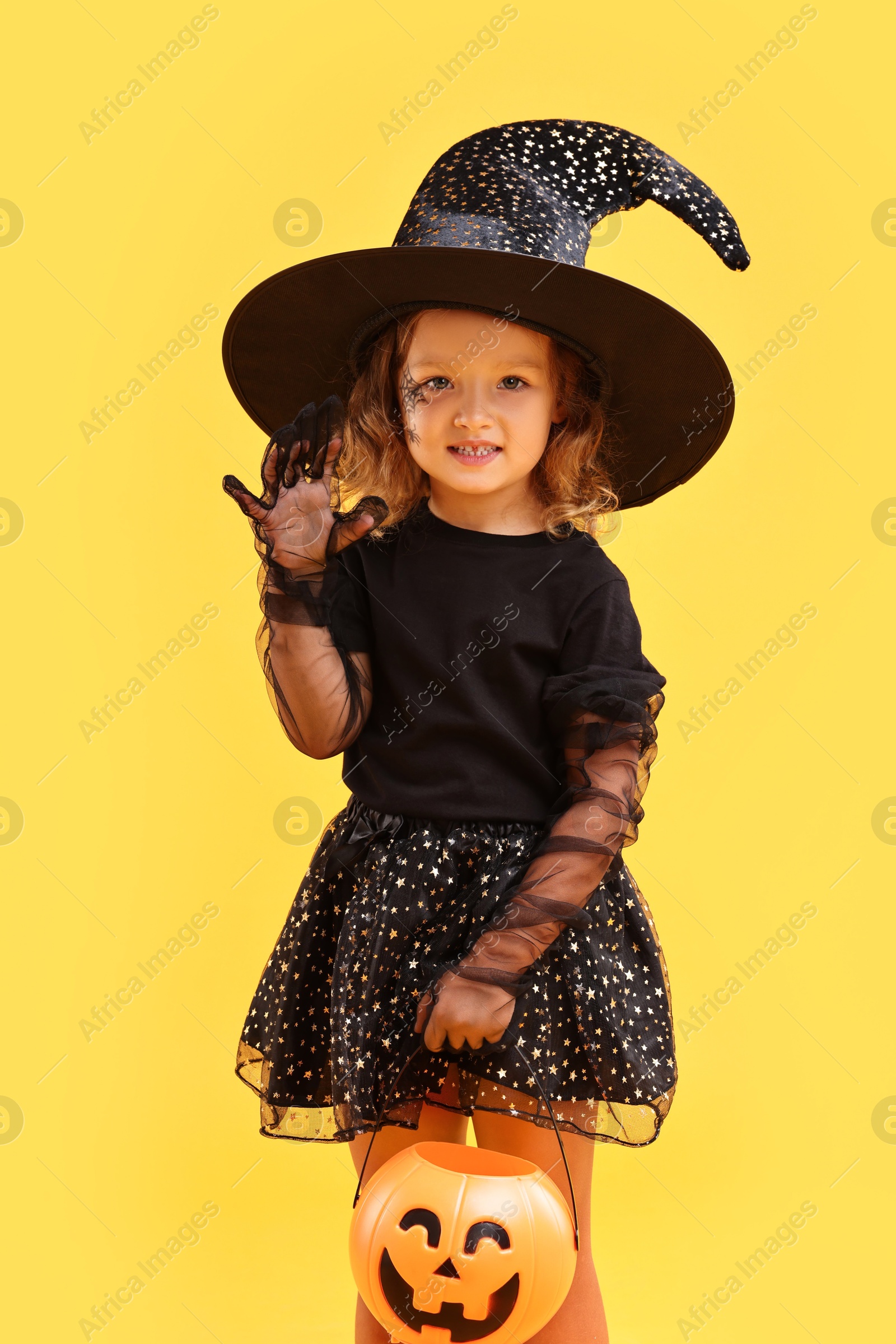 Photo of Cute girl with pumpkin bucket dressed like witch on yellow background. Halloween celebration