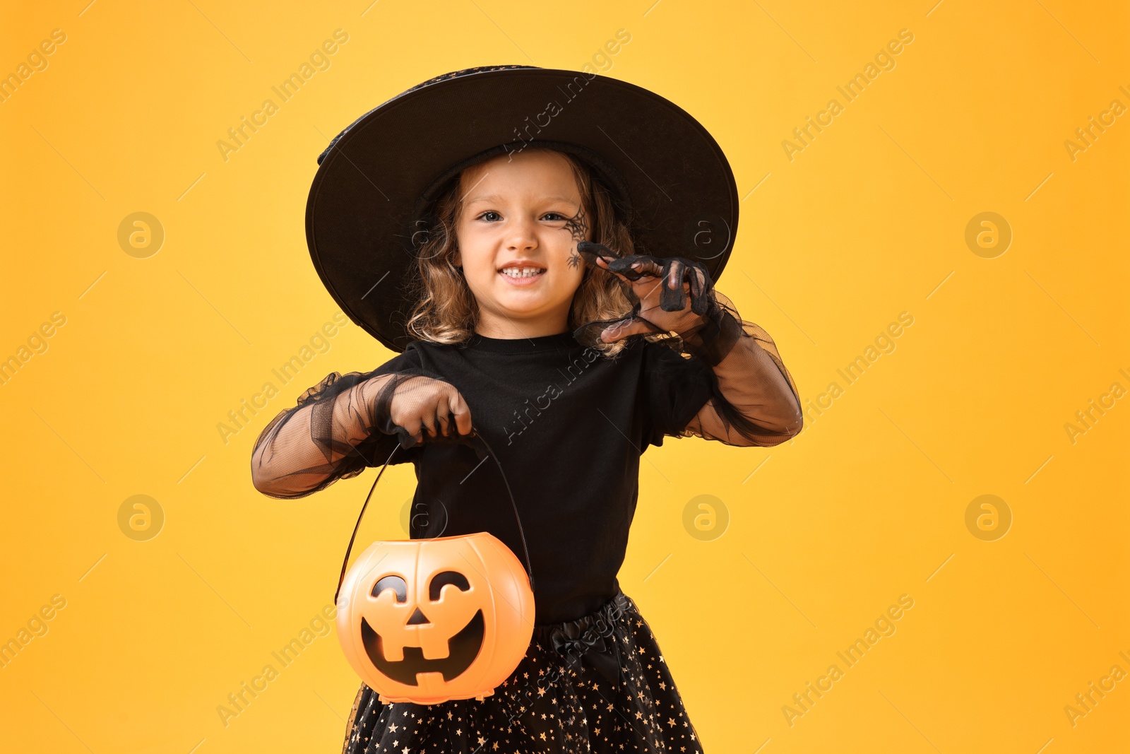 Photo of Cute girl with pumpkin bucket dressed like witch on yellow background. Halloween celebration