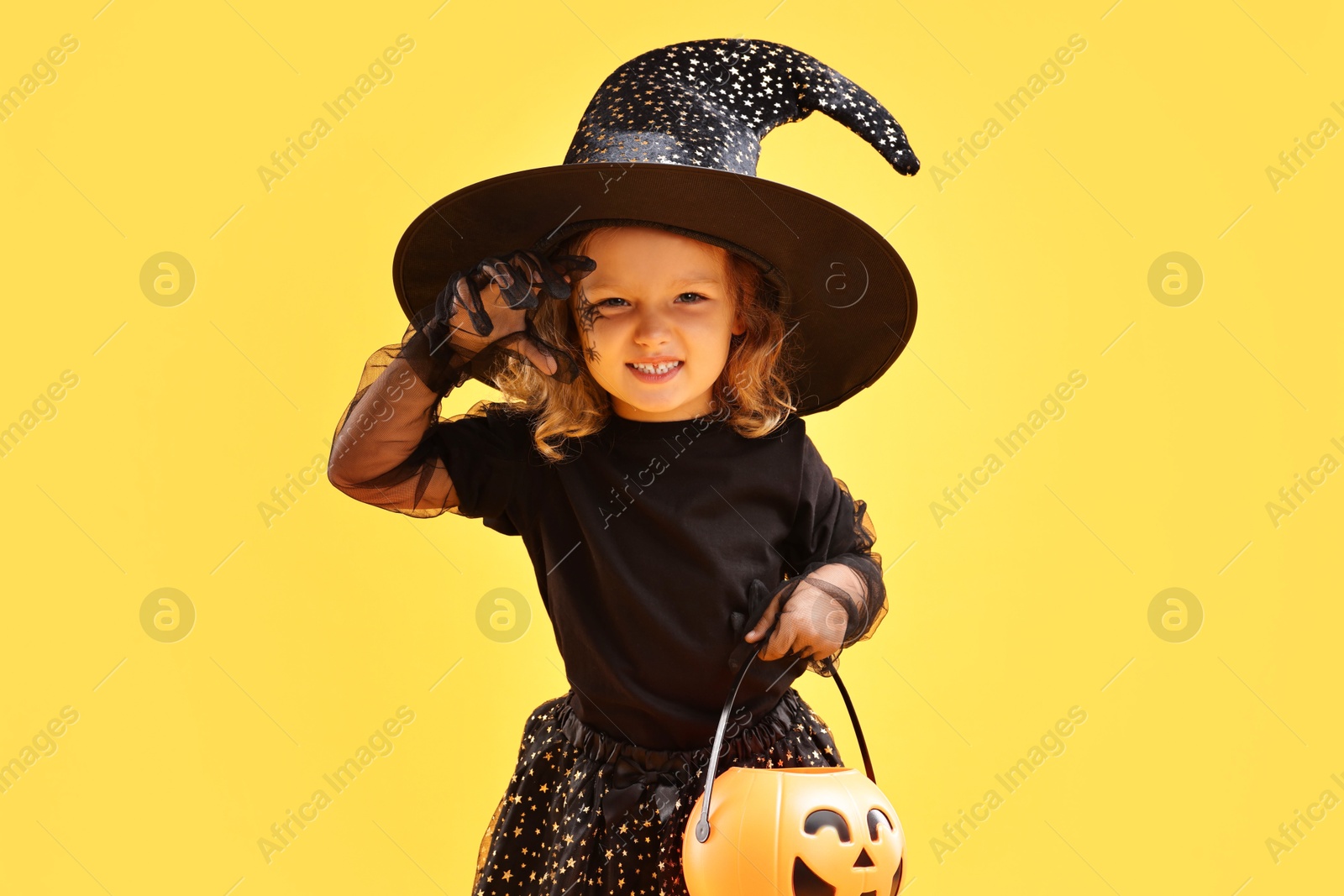 Photo of Cute girl with pumpkin bucket dressed like witch on yellow background. Halloween celebration