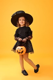 Photo of Cute girl with pumpkin bucket dressed like witch on yellow background. Halloween celebration