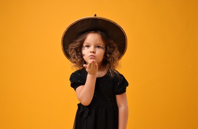 Cute girl dressed like witch blowing kiss on yellow background. Halloween celebration