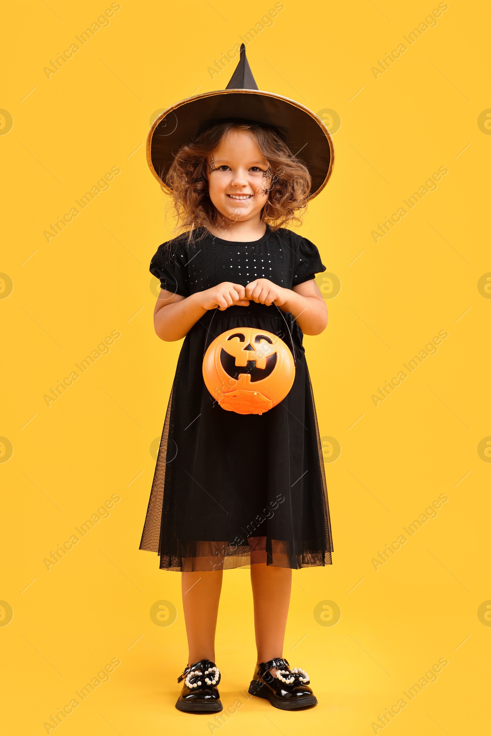 Photo of Cute girl with pumpkin bucket dressed like witch for Halloween celebration on yellow background