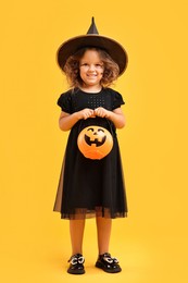 Cute girl with pumpkin bucket dressed like witch for Halloween celebration on yellow background