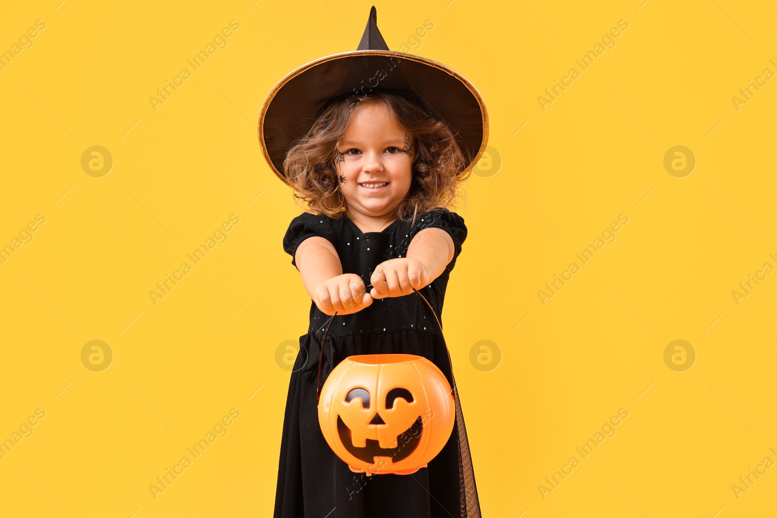 Photo of Cute girl with pumpkin bucket dressed like witch for Halloween celebration on yellow background