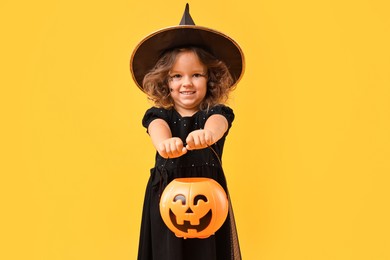 Photo of Cute girl with pumpkin bucket dressed like witch for Halloween celebration on yellow background