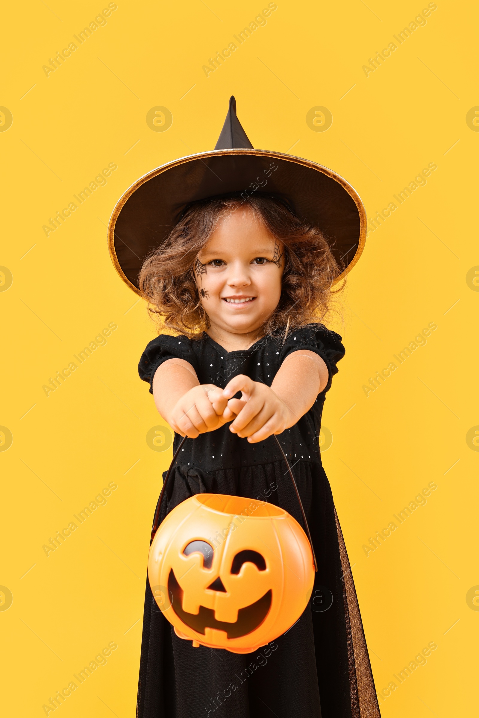 Photo of Cute girl with pumpkin bucket dressed like witch for Halloween celebration on yellow background