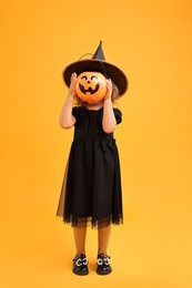 Photo of Girl with pumpkin bucket dressed like witch on yellow background. Halloween celebration