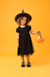 Cute girl with pumpkin bucket dressed like witch for Halloween celebration on yellow background