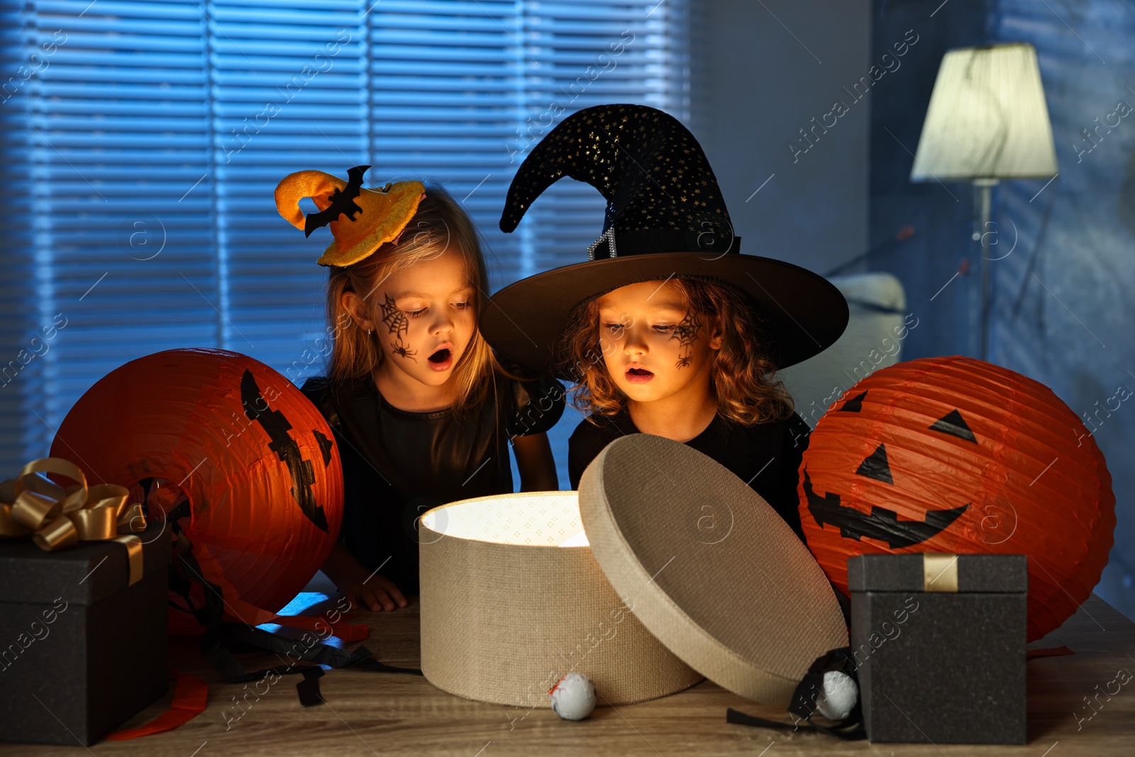 Photo of Surprised children in costumes with festive decor and gift boxes indoors at night. Halloween celebration