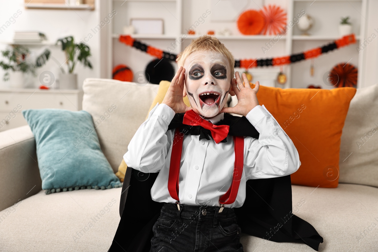 Photo of Funny boy dressed like vampire for Halloween celebration on sofa in room