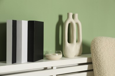 Photo of Hardcover books with different cities names, candle and vases on white shelf near green wall indoors