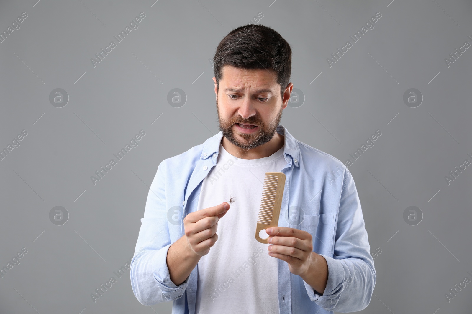 Photo of Emotional man holding clump of lost hair and comb on gray background. Alopecia problem