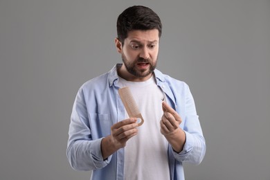 Emotional man holding clump of lost hair and comb on gray background. Alopecia problem