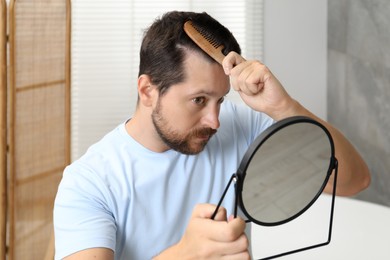 Man brushing his hair near mirror indoors. Alopecia problem