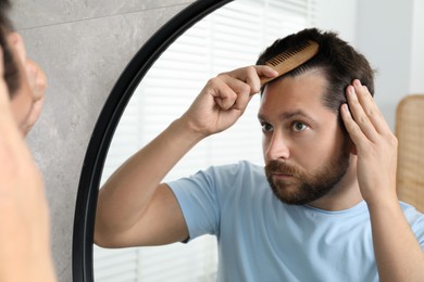 Photo of Man brushing his hair near mirror indoors. Alopecia problem