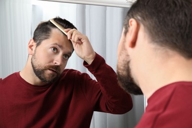 Man brushing his hair near mirror indoors. Alopecia problem