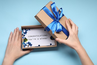 Woman opening gift box with phrase Congratulations You`re Winner on card against light blue background, top view