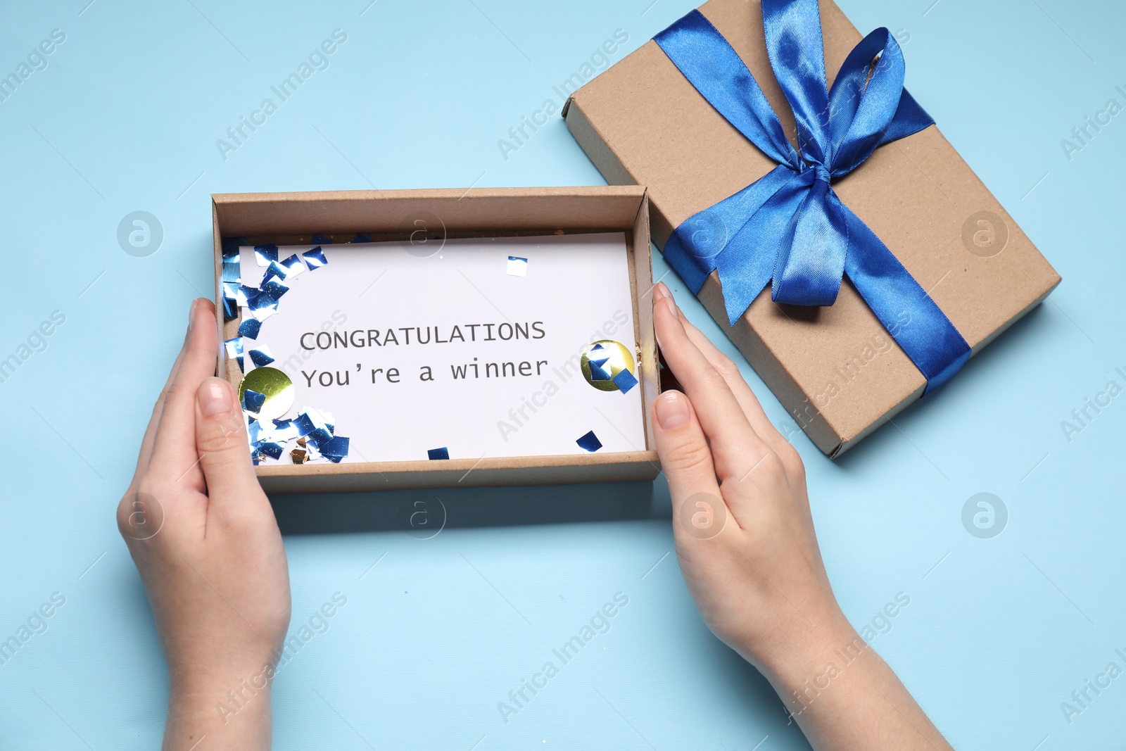 Photo of Woman opening gift box with phrase Congratulations You`re Winner on card against light blue background, top view