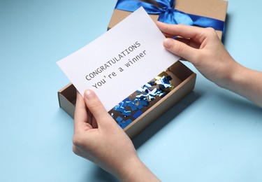 Photo of Woman holding card with phrase Congratulations You`re Winner and gift box on light blue background, closeup